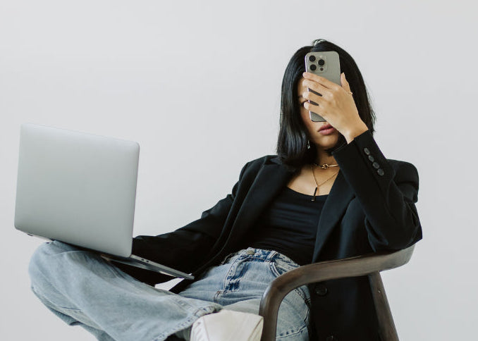 Person sitting on a chair with a laptop on their lap, holding a phone to their ear, wearing a black jacket, black top, light blue jeans, and white sneakers.